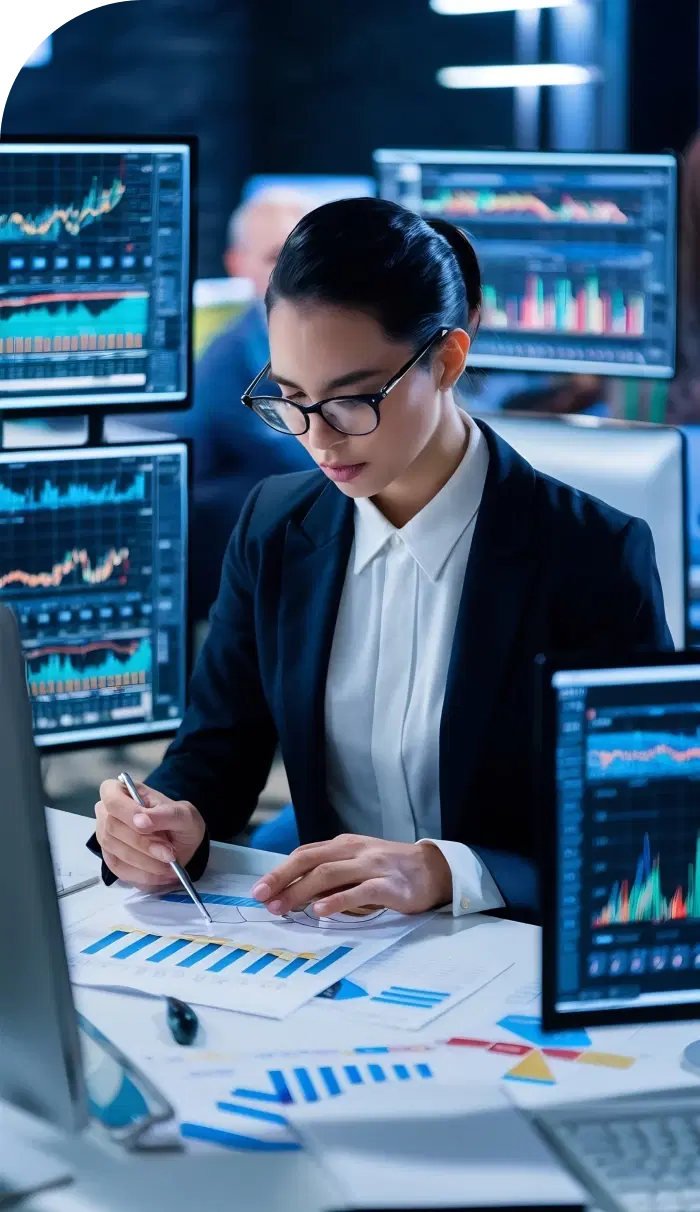 Professional reviewing financial documents at a desk with multiple monitors displaying financial data in a BFSI environment